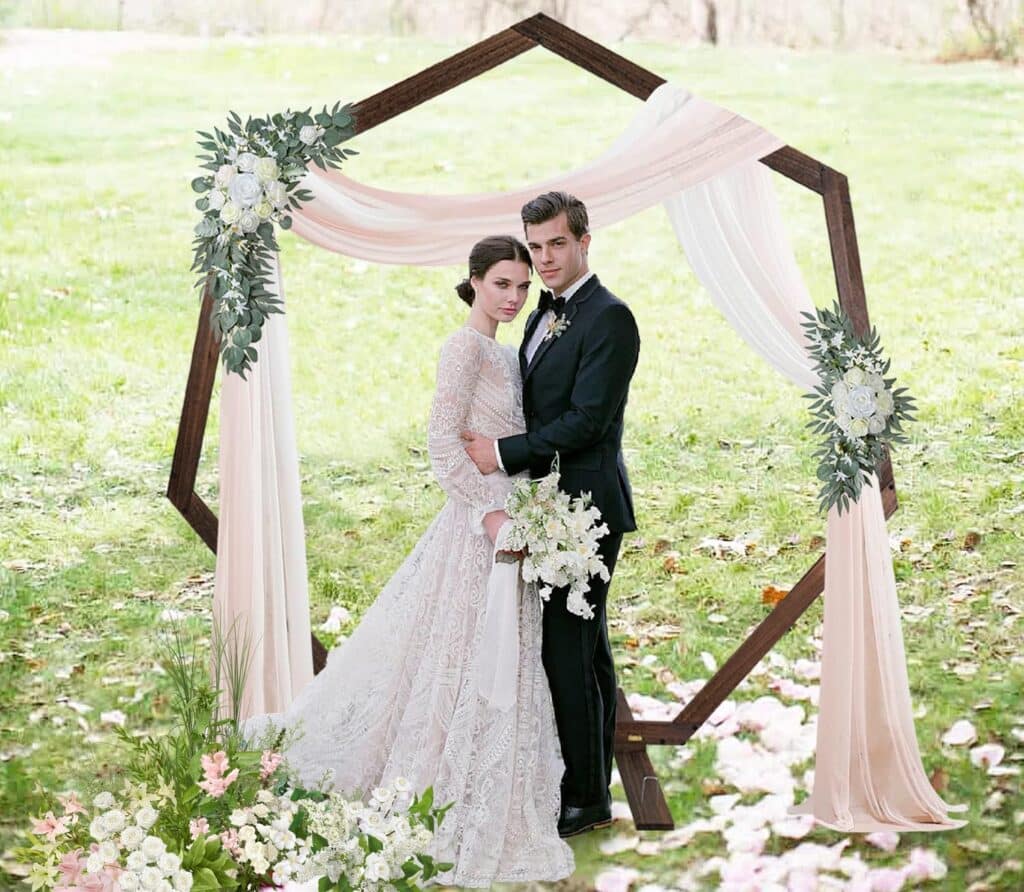 Happy Couple Share Hugs and standing next to Wooden Geometric Heptagon Arch with rose and greenery glued on